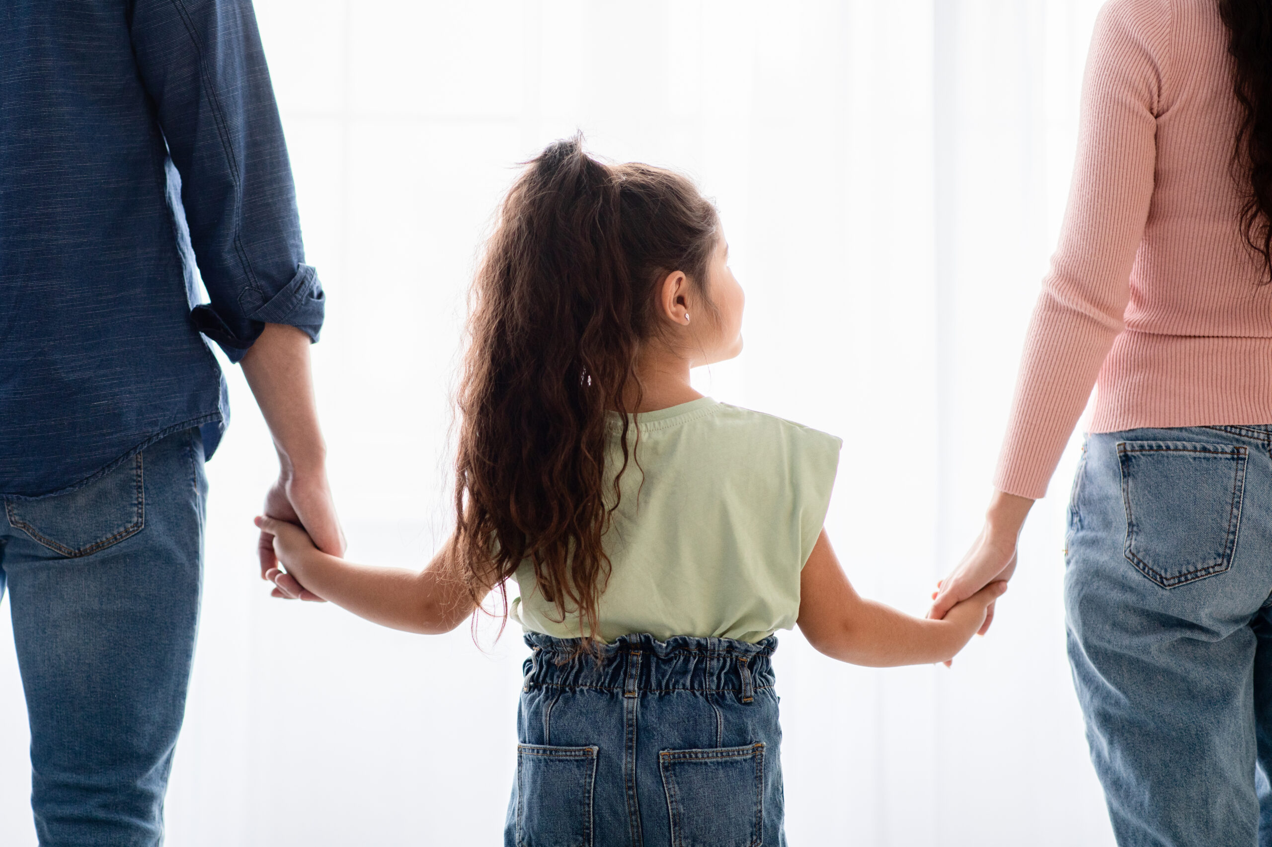 Family Support Concept. Rear View Of Little Girl Holding Hands With Mom And Dad, Standing Near Window At Home, Closeup Shot Of Parents And Female Child Bonding Together, Cropped Image
